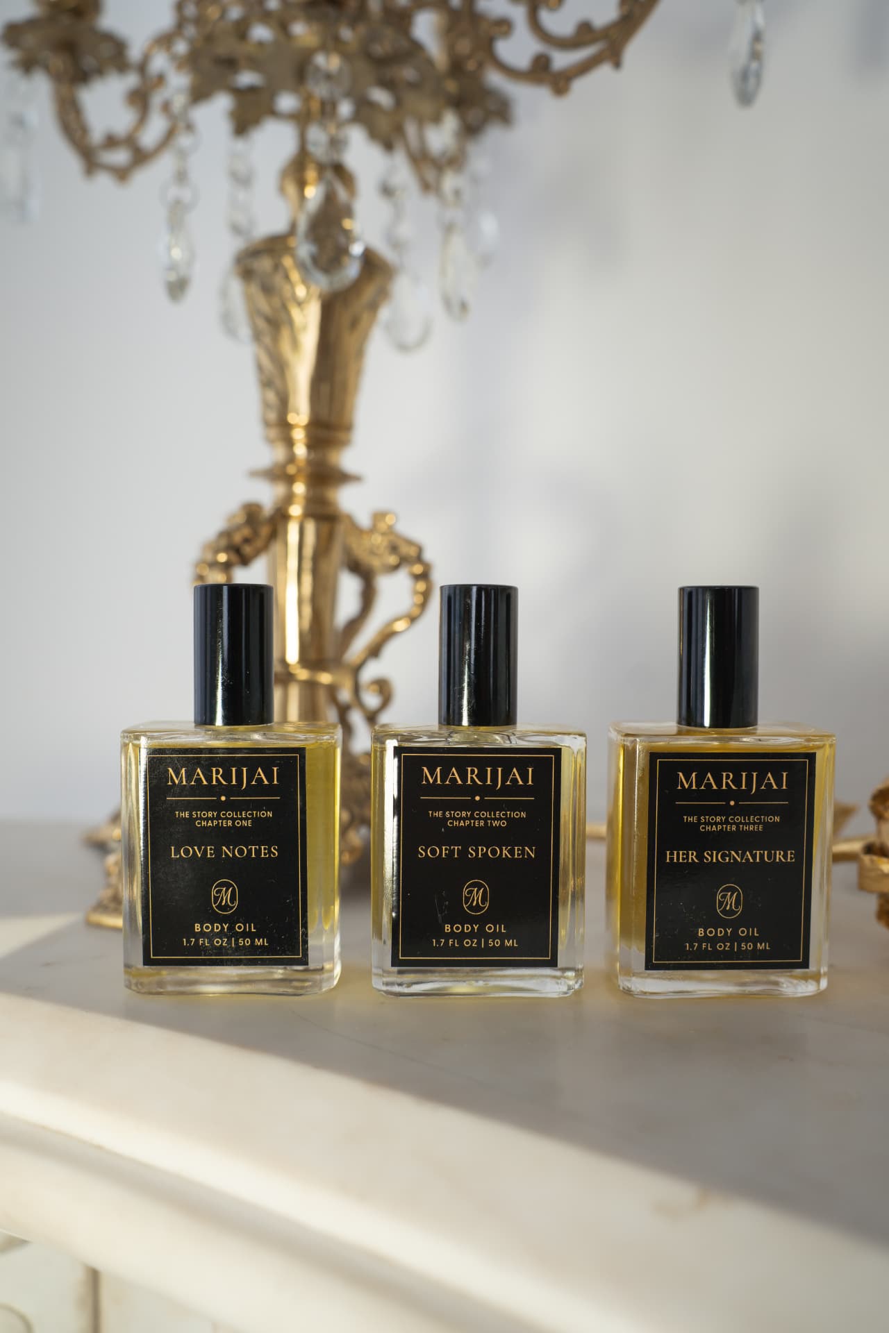 MARIJAI The Story Collection trio — Love Notes, Soft Spoken and Her Signature fragrance body oil bottles with a gold candelabra